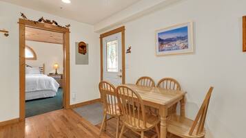 A dining area with a wooden table and chairs, a bed in the adjacent room, and a framed picture on the wall.