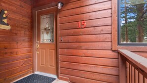 Puinen mökin sisäänkäynti, jossa on ovi, jonka yläpuolella on numero ’15’, karhukuva, joka toivottaa vieraat tervetulleiksi, ja ikkuna, josta avautuu näkymä puihin.