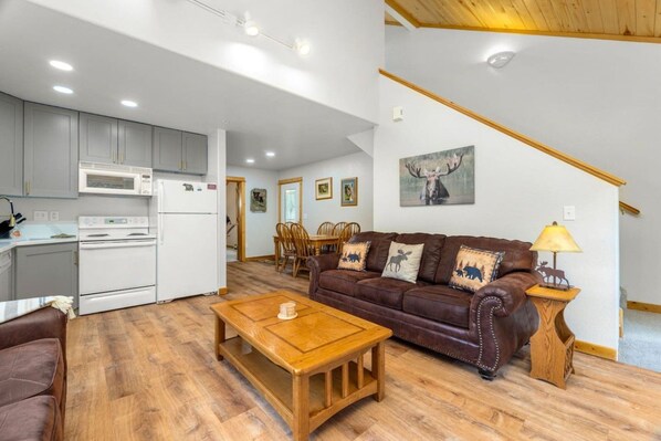 A modern kitchen with white appliances, a dining area with wooden furniture, a brown leather sofa, and a staircase with a wooden finish.