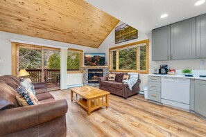 A spacious living area with a fireplace, wooden flooring, and large windows offering natural light and views of the outdoors.