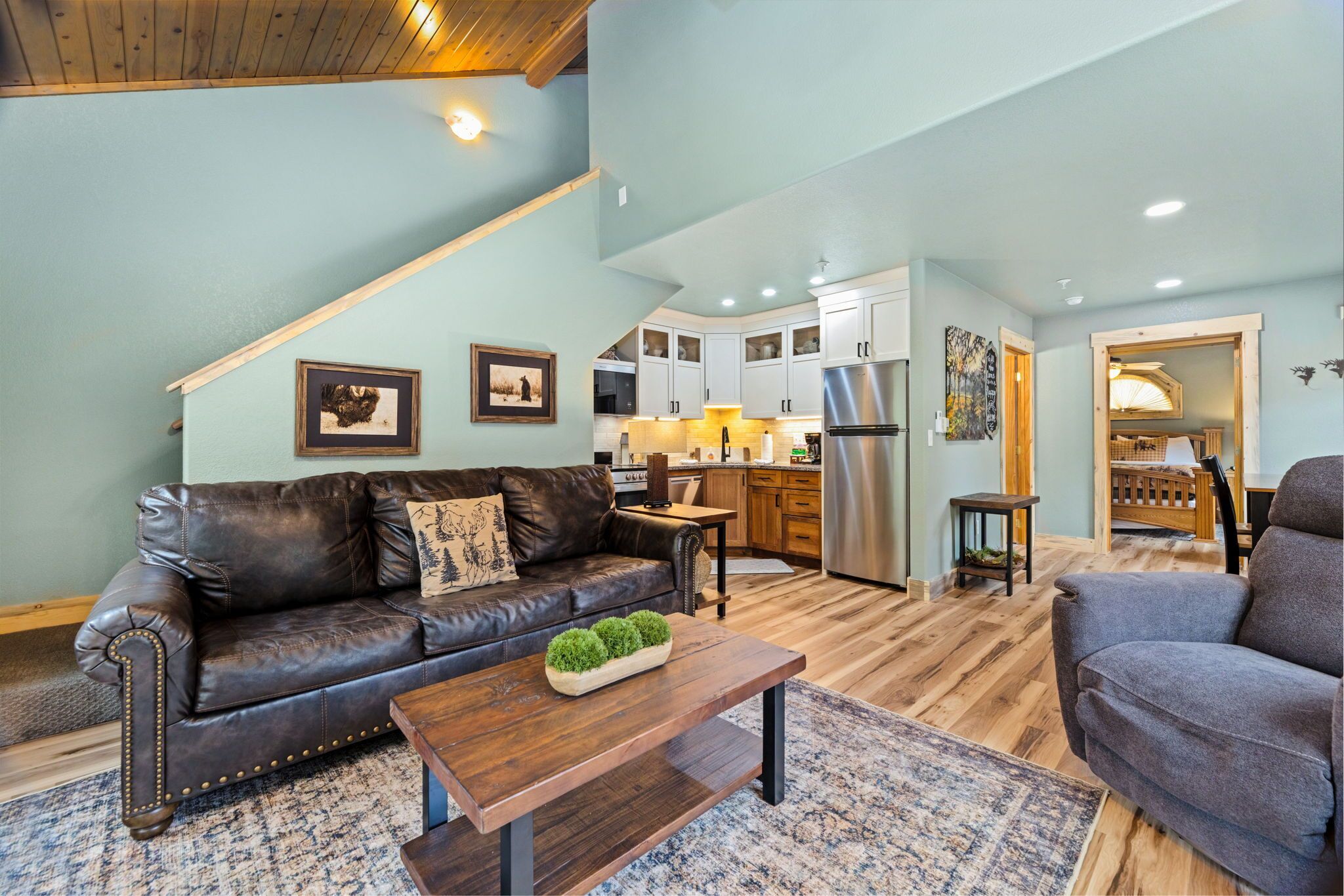 A spacious living area with a brown leather sofa, a wooden coffee table, and a gray armchair. A kitchen with white cabinets and stainless steel appliances is visible in the background.