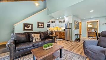 A spacious living area with a brown leather sofa, a wooden coffee table, and a gray armchair. A kitchen with white cabinets and stainless steel appliances is visible in the background.