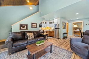 A spacious living area with a leather sofa, a wooden coffee table, and a grey armchair. A kitchen with white cabinets and a stainless steel refrigerator is visible in the background. - Riverfront Escape • A/C • Near RMNP (Estes Park)