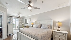 A bedroom with a large bed, two bedside tables, a ceiling fan, and a view of the outside.