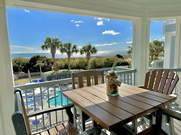 Un porche cubierto con una mesa y sillas de madera, con vistas a una piscina y una playa.