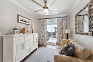A room with a ceiling fan, a white dresser with drawers, a sofa with patterned cushions, a framed picture on the wall, and a window with curtains.