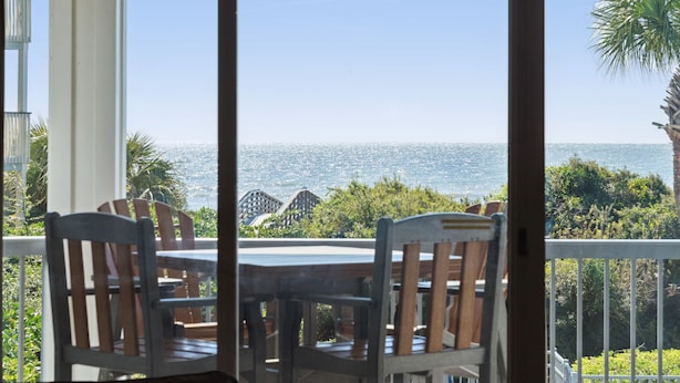 A balcony with a table and chairs, overlooking a coastal view with palm trees and buildings in the distance.