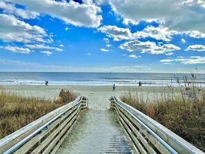 Een strand met een houten looppad dat naar het water leidt, een heldere lucht en enkele mensen die wandelen.