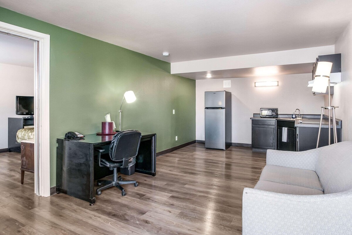 A modern living room with a green accent wall, a grey sofa, a desk with a computer, a chair, and a kitchen area with a refrigerator and microwave.