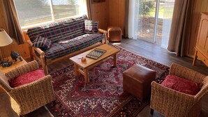 Interior acogedor de una cabaña con un sofá de cuadros, dos sillones de mimbre, una mesa de centro de madera y una alfombra estampada.