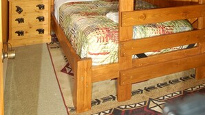 A wooden bunk bed with a patterned bedspread, a nightstand with a lamp, and a drawer unit with bear designs. - Welcome to Lakeview Pine: The Lakeside Escape (Birchwood)