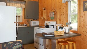 A compact kitchen with wooden walls, a refrigerator, a microwave, a stove, and a table with stools. - Welcome to Lakeview Pine: The Lakeside Escape (Birchwood)