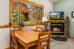 A cozy room with a fireplace, a dining table with chairs, a television, and a window with a view of a cabin and trees.