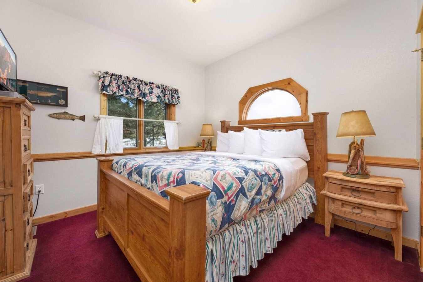 A bedroom with a wooden bed, a nightstand with a lamp, a window with curtains, and a fish decoration on the wall.