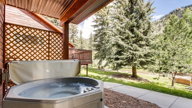 A hot tub in a covered outdoor area with a wooden deck and a view of a forested hillside. - Riverfront Hideaway • Private Hot Tub• Close to RMNP (Estes Park)