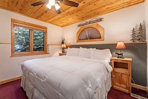 A bedroom with a large bed, a ceiling fan, a window with a view of trees, a wall-mounted sign, and a nightstand with a lamp. - Fall River Hideaway • Private Hot Tub (Estes Park)
