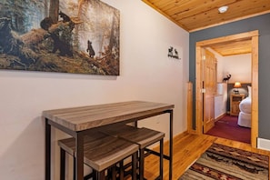 A hallway with a rustic wooden table and chairs, a painting of a forest scene, and a door leading to a bedroom with a bed and a nightstand.