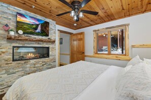 A bedroom with a stone fireplace, a television mounted on the wall, and a window with a view of the outside. - Renovated Fall River Escape • Private Hot Tub (Estes Park)