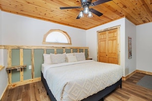 Une chambre avec un plafond en bois, un lit recouvert d’une courtepointe blanche, une tête de lit en bois et une fenêtre avec un cadre en bois.