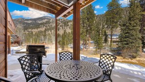 A covered patio with a round table and chairs, overlooking a snowy mountain landscape. - Riverfront Escape •Private Hot Tub • Close to RMNP (Estes Park)