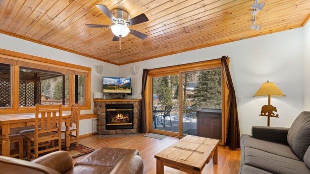 A cozy living room with a fireplace, wooden furniture, and large windows offering a view of the outdoors. - Riverfront Escape •Private Hot Tub • Close to RMNP (Estes Park)
