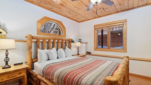 A wooden-framed bed with a striped bedspread, a nightstand with a lamp, a ceiling fan, and a window with a view of the outside. - Riverfront Escape •Private Hot Tub • Close to RMNP (Estes Park)