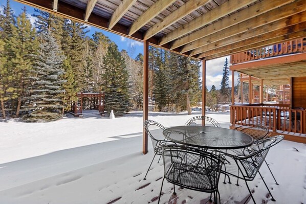 A covered outdoor seating area with a table and chairs, surrounded by snow and trees.