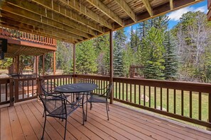 A covered outdoor deck with a table and chairs, surrounded by a wooden railing, overlooking a forested area.
