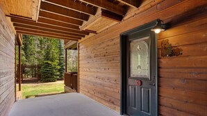 Puinen mökin sisäänkäynti, jossa on tumma ovi, pieni kuisti ja näkymä metsäiselle alueelle.