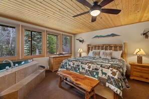 Ein Schlafzimmer mit einem großen Bett, einer hölzernen Bank, einem Nachttisch, einer Lampe, einem an der Wand befestigten Regal und einem Fenster mit Blick auf Bäume.