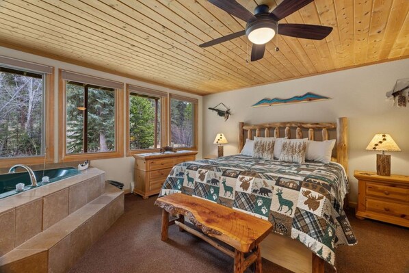 A bedroom with a large bed, a wooden bench, a nightstand, a lamp, a wall-mounted shelf, and a window with a view of trees.
