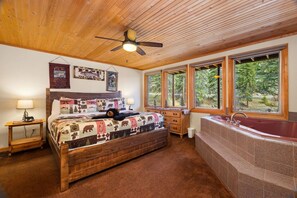 A bedroom with a large bed, a wooden ceiling fan, a window with a view of trees, a wooden dresser, and a bathtub.