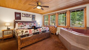 Une chambre avec un plafond en bois, un grand lit recouvert d’une courtepointe, une table de chevet, une commode, une fenêtre donnant sur des arbres et une baignoire.