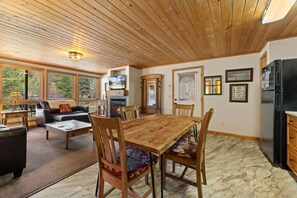 A spacious living area with a wooden ceiling, a fireplace, a dining table with chairs, and a kitchen area with a refrigerator.