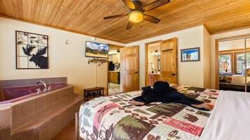 A bedroom with a large bed, a wooden ceiling fan, a bathtub, and a wall-mounted TV.
