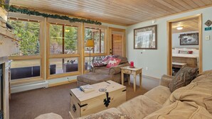 A cozy cabin interior with a wooden ceiling, large windows, a sofa, a table, and a chest of drawers. - Quiet Fall River Suite • River Views • RMNP (Estes Park)