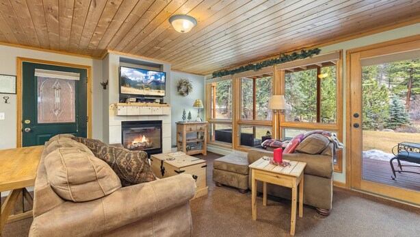 A cozy living room with a fireplace, wooden ceiling, and large windows overlooking a deck. - Quiet Fall River Suite • River Views • RMNP (Estes Park)