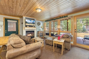 A cozy living room with a fireplace, comfortable seating, and large windows offering a view of the outdoors.