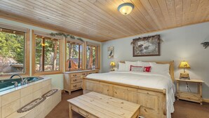 A wooden cabin bedroom with a large bed, a wooden bench, a sink, and a window with a view of trees. - Quiet Fall River Suite • River Views • RMNP (Estes Park)
