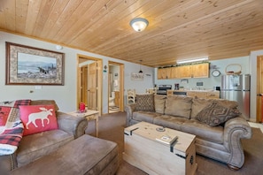 A cozy living room with a sofa, ottoman, and a coffee table. There is a kitchen area with wooden cabinets and a refrigerator.