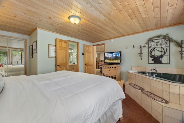 A bedroom with a large bed, a wooden ceiling, a television, and a bathroom with a sink and mirror.
