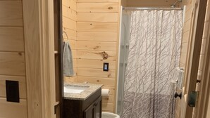 Baño de cabaña de madera con suelo de baldosas de piedra, inodoro, lavamanos con espejo y ducha con cortina estampada.