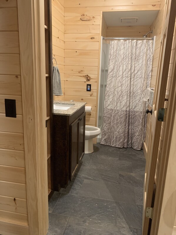 Een houten badkamer met een stenen vloertegel, toilet, een wastafel met spiegel en een douche met een decoratief gordijn.