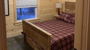 Un dormitorio de cabaña de madera con cama, mesita de noche, ventana con persianas y una lámpara en el techo.