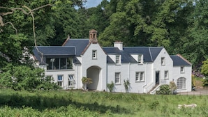 Une grande maison blanche à deux étages, dotée d’un toit à forte pente, d’une arche centrale imposante et d’une petite structure en forme de tour sur le toit.