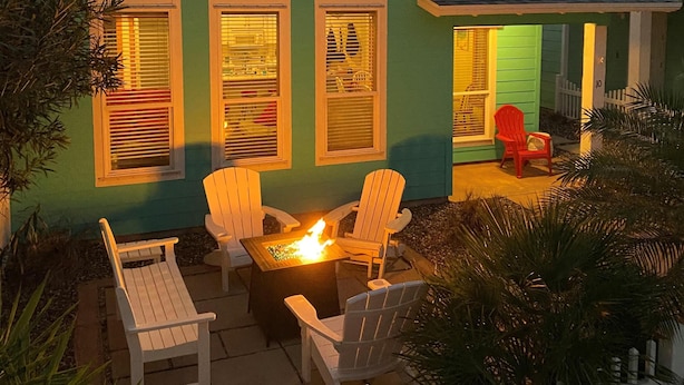 Een huis met een groene gevel, een veranda met witte leuningen en een vuurkorf met stoelen.