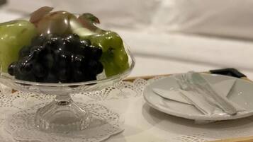 A dessert tray with a glass bowl containing mixed fruits on a doily, a plate with a fork, and a bed with white bedding in the background.