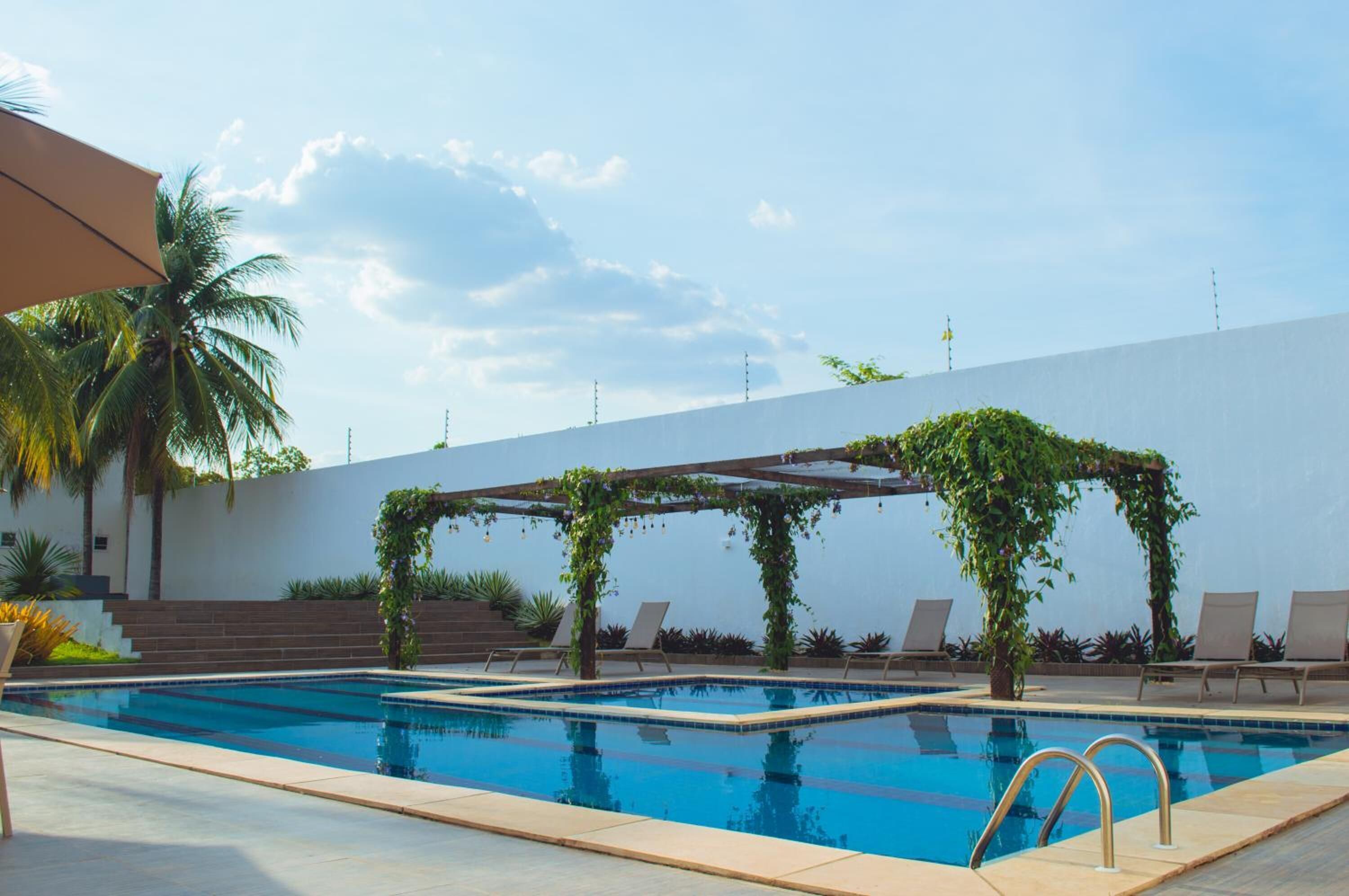 Una piscina con una pérgola cubierta de follaje, sillas de descanso y una palmera.
