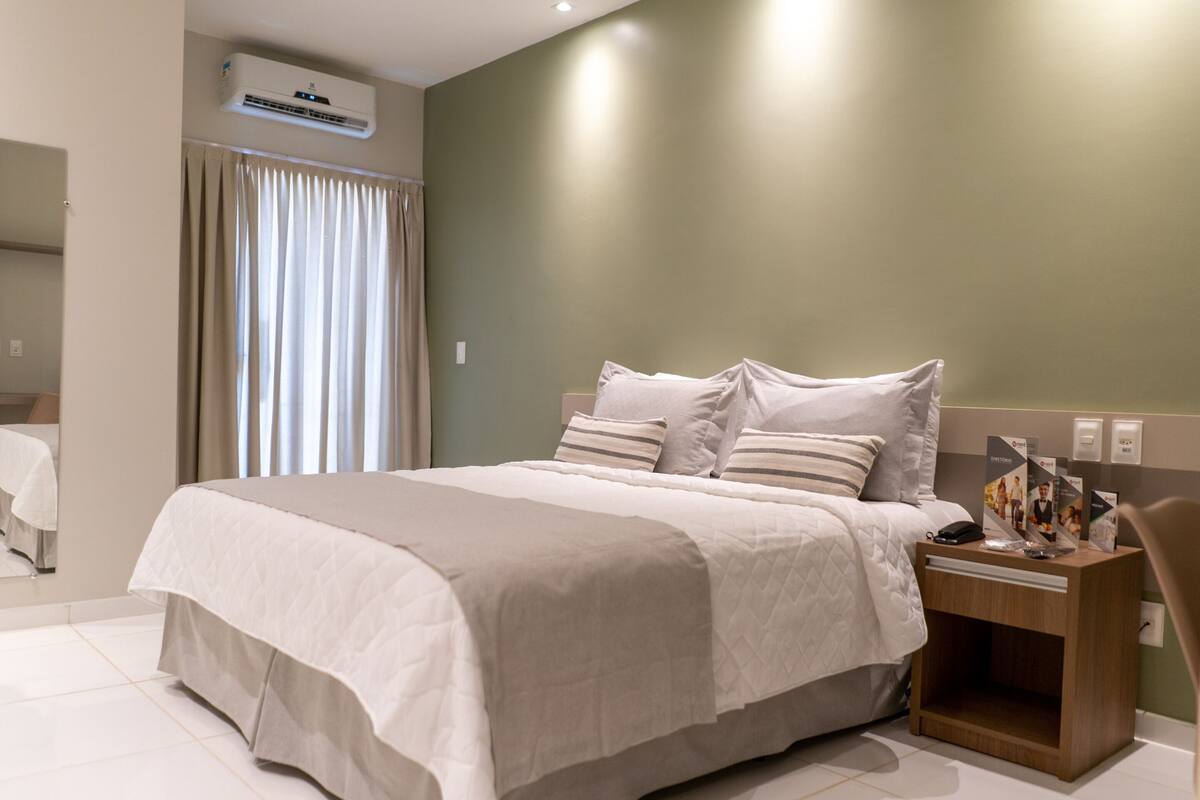 A modern hotel room with a large bed, a wooden nightstand, and a built-in air conditioning unit.