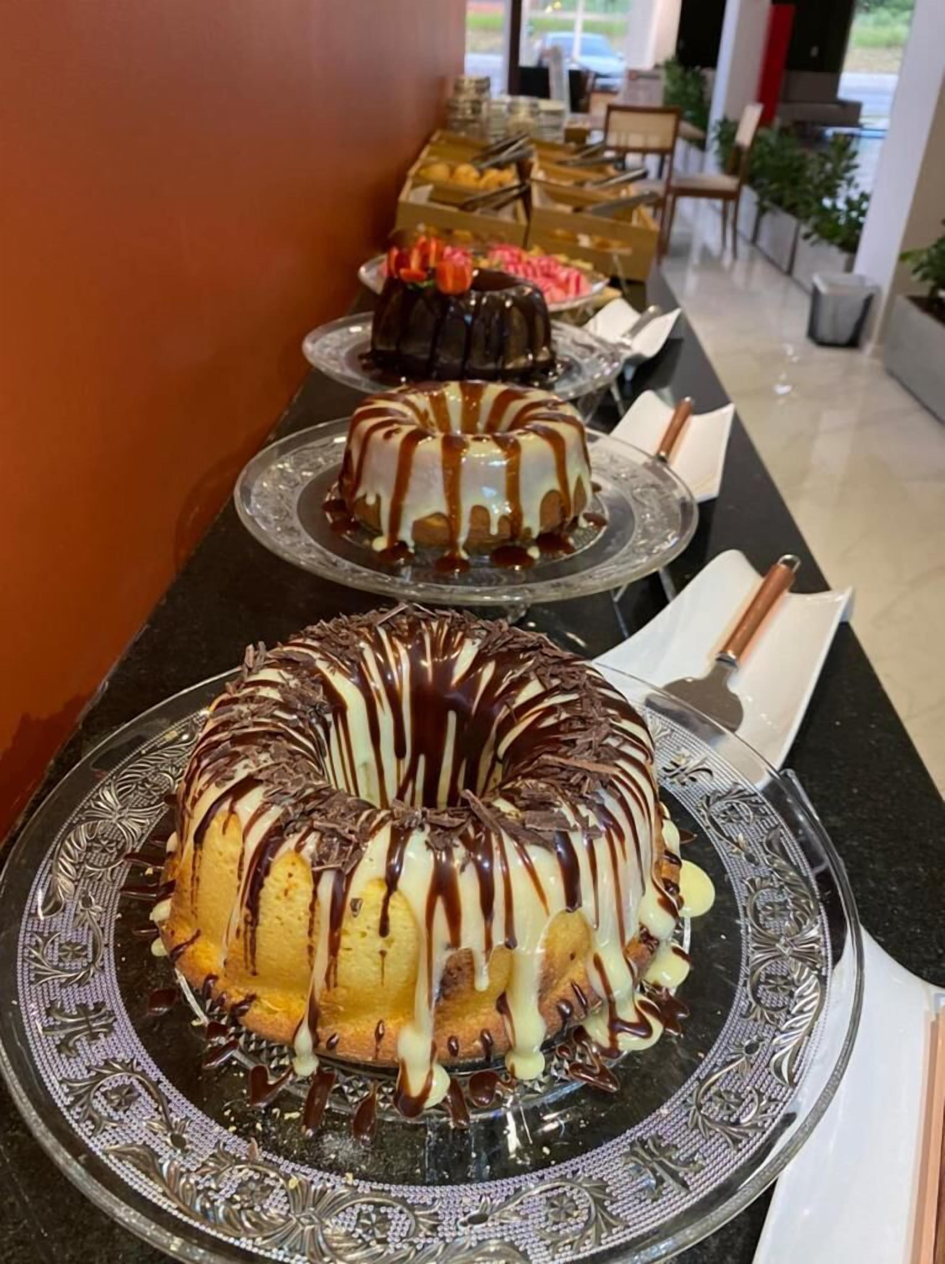 A buffet table with various desserts, including a bundt cake with chocolate drizzle and a cake with white icing and chocolate shavings.